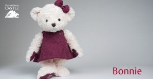 A photograph of a white bear toy wearing a dark red dress and a dark red bow.