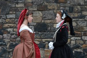 A photograph of two women dressed in Elizabethan dresses, looking at one another.