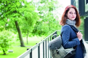 woman with red hair holds tweed backpack and wears tweed scarf as she looks at the camera