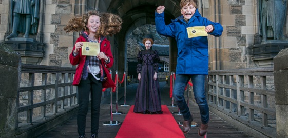 two children outside a castle jump for joy as a renaissance queen looks on in the background