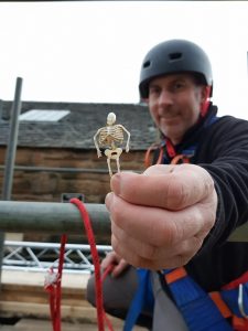 A man in a hard hat holds up a tiny plastic skeleton to the camera