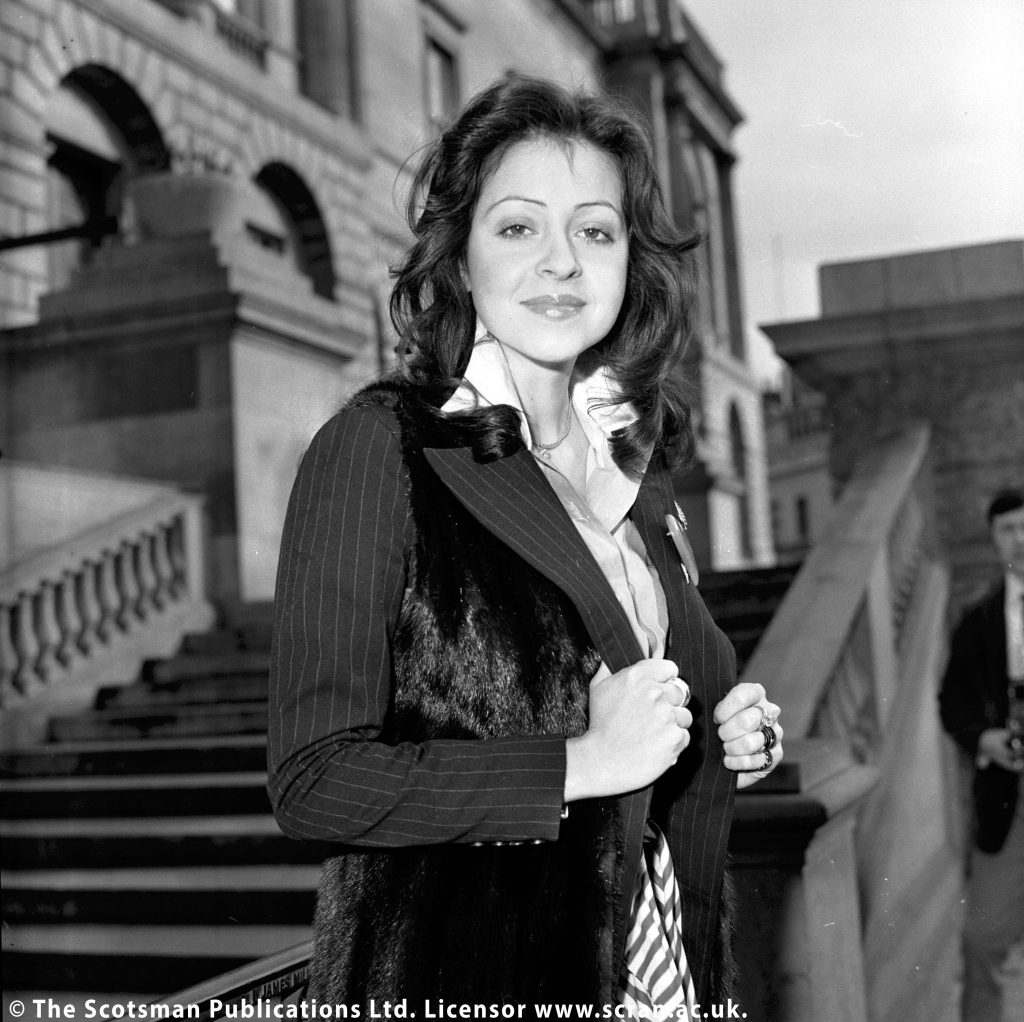 Vicky Leandros poses for a press shot in Edinburgh. She has long black hair and wears a black jacket and white shirt. The jacket features a pinstripe design. 