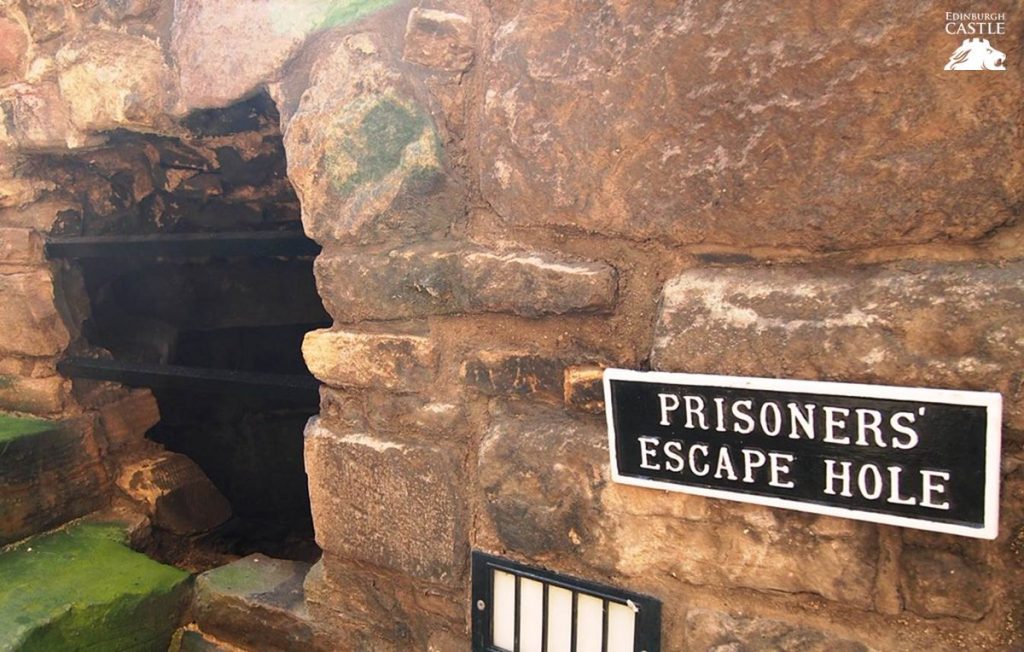 A hole in a ancient brick wall. A black and white sign beside the hole reads "Prisoners' Escape Hole." 