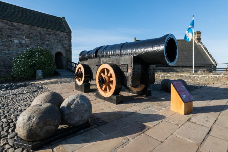 A large and ancient black cannon resting on a wooden frame with wooden and metal wheels. Three cannon balls have been placed nearby and a Scottish saltire flies from a white flag pole in the background.