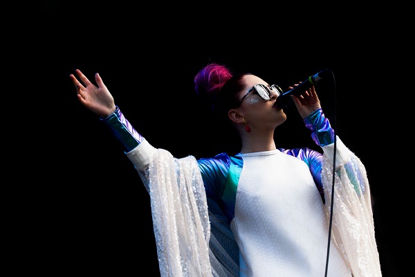 An photo of Be Charlotte. She is wearing a white and purple outfit with sunglasses. She is singing into a microphone and holding her arm aloft. 