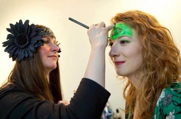 A lady wearing a black fascinator in her hair applies face make-up to another lady. Image credit Ruth Armstrong