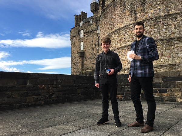 Two magicians pose outside Edinburgh Castle. Both are wearing tartan shirts. One holds a rubix cube and the other a deck of playing cards. 