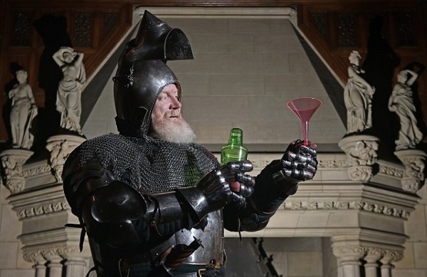 A bearded man dressed in armour and chain-mail prepares a cocktail. In his left hand he is holding a green bottle and in his right hand he is holding a pink cocktail glass. He is standing in front of an ornate stone fireplace. 