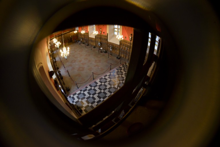 A view across the Great Hall of Edinburgh Castle from the Laird's Lug. The entire room can be seen which includes wood paneling, suits of armour and chandeliers. Weaponry hangs on the walls and the floor features intricate tiling patterns. 