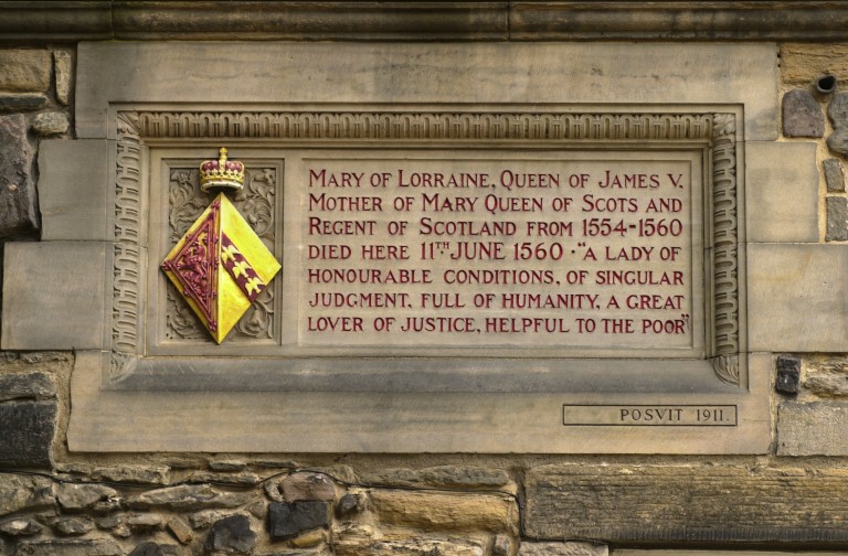 A plaque built into a castle wall. Red lettering commemorates the death of Mary of Lorraine in 1560. A red and yellow coat of arms topped with a crown is next to the text.