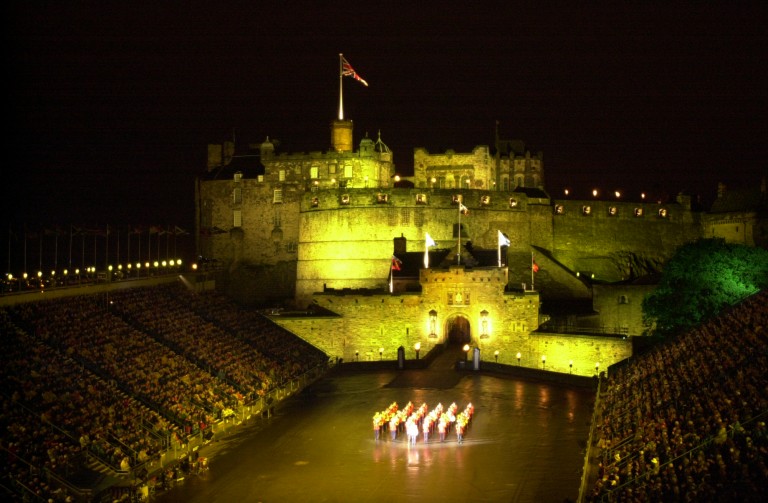 A scene from the Royal Edinburgh Military Tattoo. A military band is performing on the Castle Esplanade which is flanked by seated spectators. The Castle is floodlit in the background. 