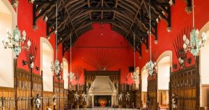 The Great Hall at Edinburgh Castle. The walls are red with a high timbered ceiling. Weapons and armour hang on the walls and a grand stone fireplace is at the end of the room.