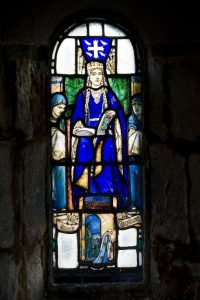 A stained glass depiction of St Margaret wearing blue robes and gold crown. An open book, perhaps a Bible, is on her lap.