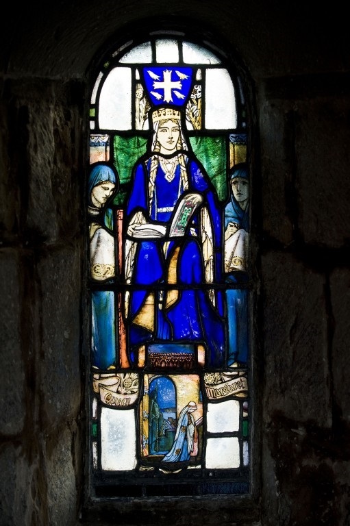 A stained glass depiction of St Margaret wearing blue robes and gold crown. An open book, perhaps a Bible, is on her lap. 