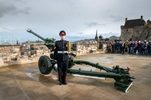 A woman in uniform stands to attention in front of a field gun. THe Edinburgh skyline is behind her