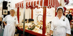 Two women in aprons and mop caps stand in front of a small market stall laden with sweet treats