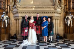 A wedding party pose for a photo in front of a large stone fireplace inside Edinburgh Castle