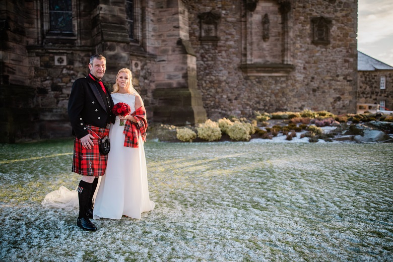 A woman in a wedding dress and a man in a kilt pose for a photo in the grounds of Edinburgh Castle. There is an attractive layer of frost on the grass. 