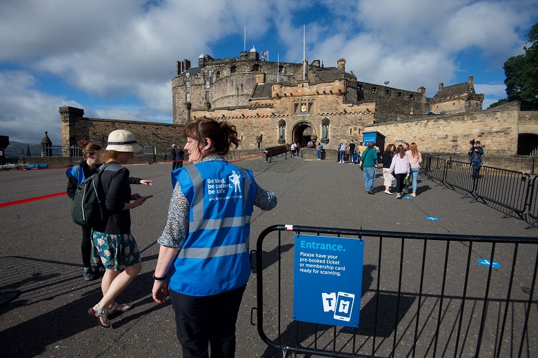 A member of staff standing beside a sign marking the entrance to Edinburgh Castle. A socially distanced queue of visitors is forming in the background.