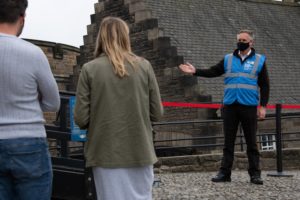 A member of staff in a blue jacket and a black face mask welcomes two visitors to Edinburgh Castle