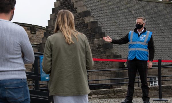 A member of staff in a blue jacket and a black face mask welcomes two visitors to Edinburgh Castle