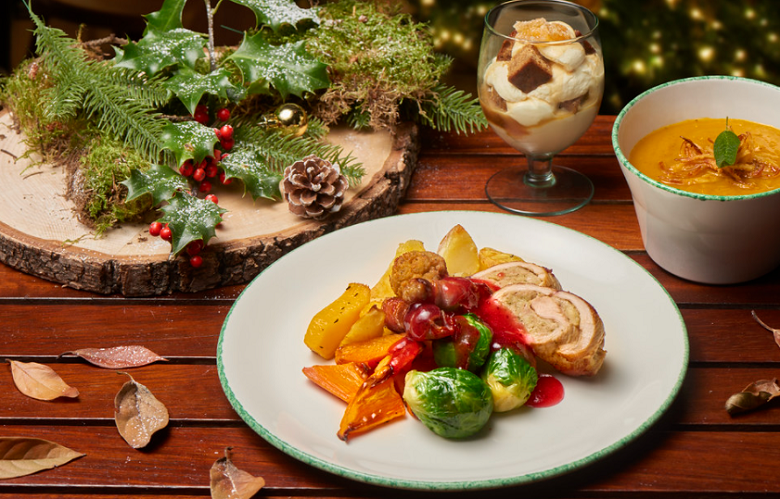 A festive-themed meal on a table adorned with rustic decorations including holly. The food includes soup, roast turkey, brussels sprouts and a decadant chocloate-based dessert served in a tall glass.