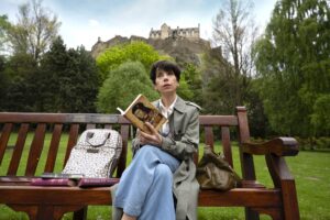 Film still from "The Lost King" showing Sally Hawkins siting on bench in front of Edinburgh Castle with books about King Richard III