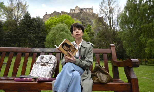 Film still from "The Lost King" showing Sally Hawkins siting on bench in front of Edinburgh Castle with books about King Richard III