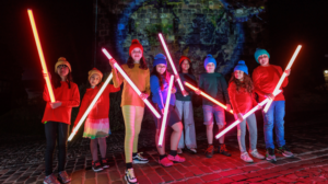 A group of young people at Edinburgh Castle of Light holding glowing sticks standing in front of a light installation.