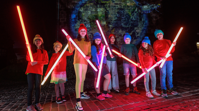A group of young people at Edinburgh Castle of Light holding glowing sticks standing in front of a light installation.