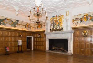 A large room with a grand stone fireplace. There is wood paneling on the walls and above that painted frescoes. The ceiling is made of ornate panels too.
