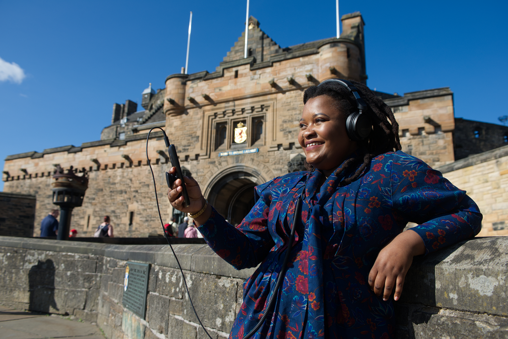 The artist, Tanatsei Gambura, on Edinburgh Castle esplanade wearing headphones and smiling looking at her mobile phone