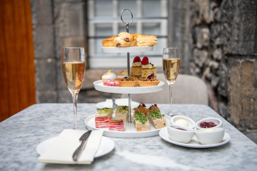 An etagere of afternoon tea with sandwiches and cakes and scones. Beside it are scoops of jams and clotted cream and two glasses of fizz