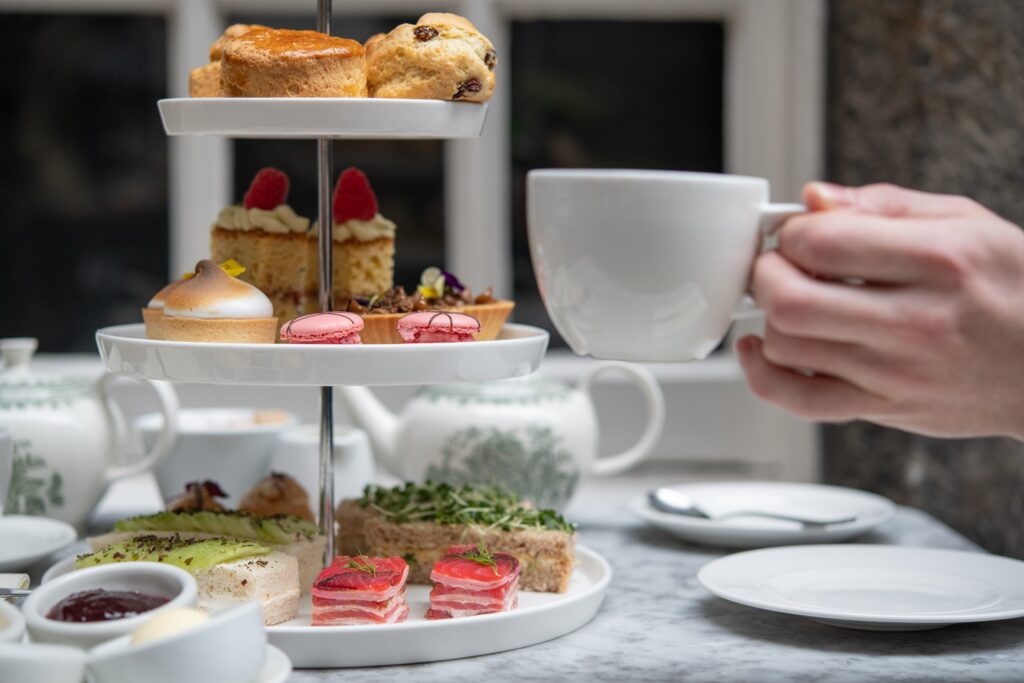 Close up of a hand holding a white teacup. In the background you can see an afternoon tea etagere. 