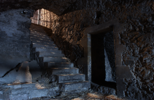 dimly lit stone staircase leads upward to a barred gate or window, where light streams in. The steps are worn and uneven, surrounded by rough stone walls. A dark passageway opens to the right, and the floor is scattered with straw, creating a mysterious, aged atmosphere.